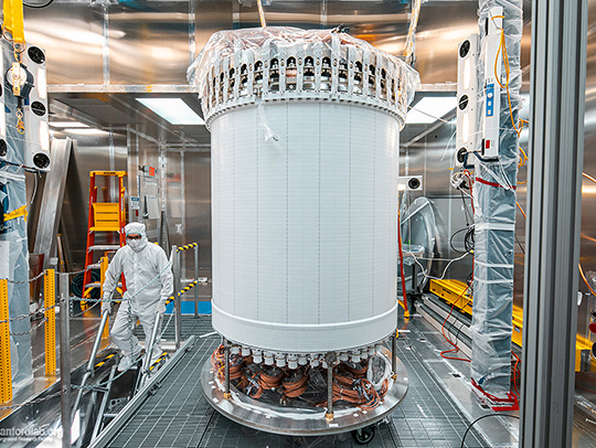 A scientist in a clean suit goes down a set of steps next to a very large, cylindrical white piece of equipment in the Sanford Underground Research Facility. Credit: Matthew Kapust, Sanford Underground Research Facility.